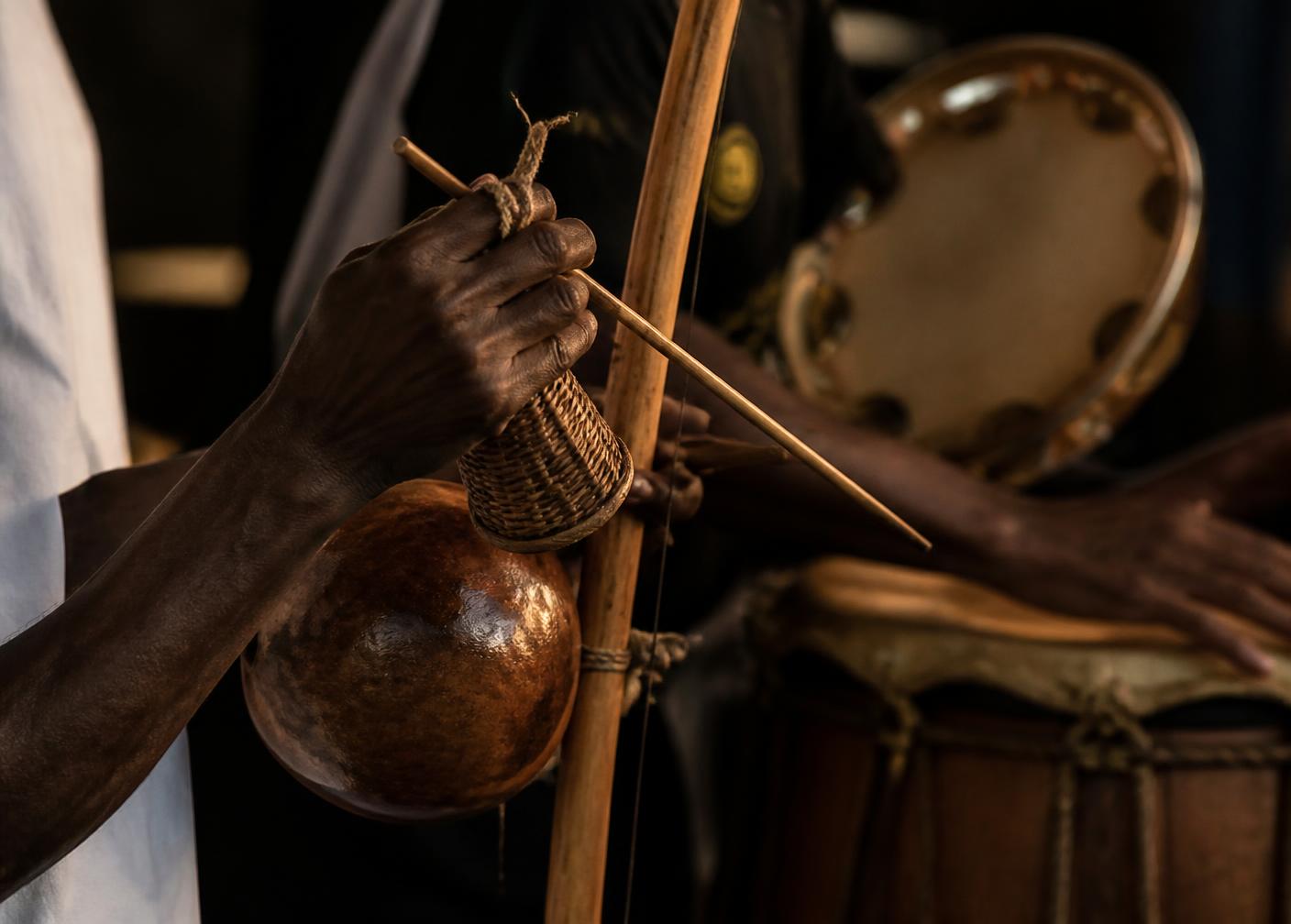Mãos tocando berimbau ao lado de atabaque e pandeiro